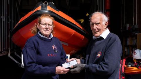 Liz Wilson, left, stands in front of an orange RNLI vessel, as she and Neil Anderson, right, hold up an award. 