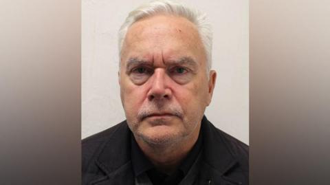 A custody picture of Huw Edwards. He has short, grey hair with stubble and is wearing a black top and jacket.