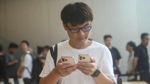 Young man is shown wearing a white t-shirt, holding an iPhone 16 model in each hand, with a thoughtful expression on his face at an Apple Store in Hangzhou, China.