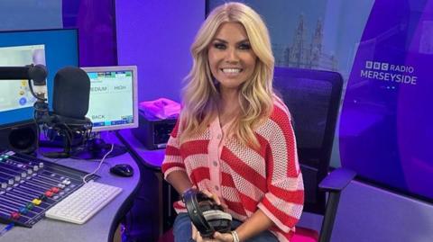 Leanne Campbell, who has wavy blonde hair and is wearing a chunky knit red and white striped cardigan, sits in a swivel chair by a studio desk with a large presenter's microphone to her right. She is holding a pair of black earphones.