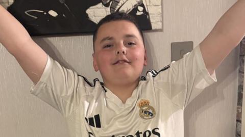 A boy with dark hair and a white football shirt on with both arms raised in the air
