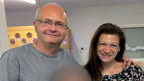 Maxie Allen (right) and his partner Rosalind Levine smile while posing for a photo and looking directly at the camera standing in what looks like some kind of locker room.