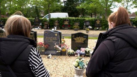 Two women stand with their backs to the camera, looking on at the grave of a loved one. Two gravestones stand before them, decorated with floral tributes. A row of stones sit behind that, then a hedge and a road behind it.