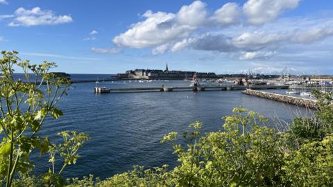 A wide view of working harbour on a sunny day, with a medieval city in the background