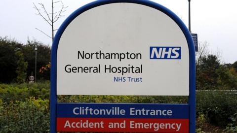 A white and blue sign next to bushes at the hospital. It says Northampton General Hospital NHS Trust, then below: Cliftonville Entrance, and in a red strip below that: Accident and Emergency.