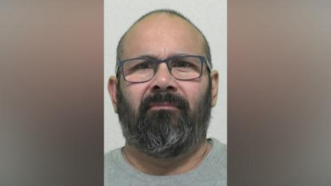 Mugshot of Hussain. He has balding short dark hair and a thick black and grey beard. He is wearing glasses and a grey t-shirt.