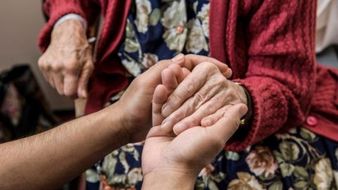 An elderly woman in a red cardigan and patterned dress has her hand being held in the palms of a younger person. Her face is not visible.