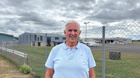Former Wing Commander Simon Footer smiling at the camera. He has grey hair and is wearing a light blue polo shirt. Behind him is a metal fence and a plane and buildings at an RAF base. 