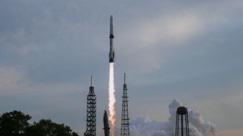 Blue Origin's New Glenn rocket taking off from a Florida launchpad with a hazy blue sky behind it.