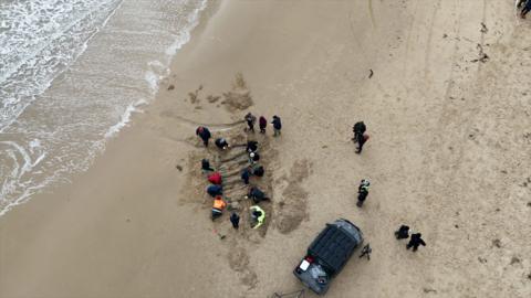 An aerial view of people gathered around the outline of part of the ship in the sand. There is a car parked nearby and some whitewash from the waves can be seen to the left.