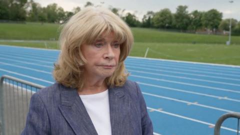 A woman with blonde hair wearing a white t-shirt and grey blazer stands in front of a blue running track