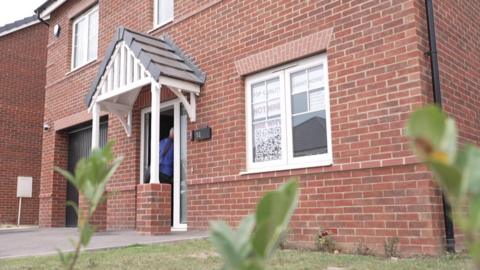 A newbuild red-brick detached house. It has white window frames. In the window closest to the camera are signs reading: Top quality Avant Homes not mine See why...
