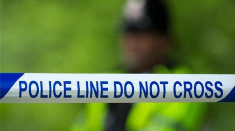 A police officer in a high vis jacket and black hat can be seen, out of focus, behind blue and white police tape