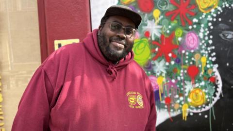 A man wearing a red hooded top with a baseball cap, beard and glasses is stood in front of a boarded up window which has been painted with the image of a lady with bright coloured afro hairstyle 