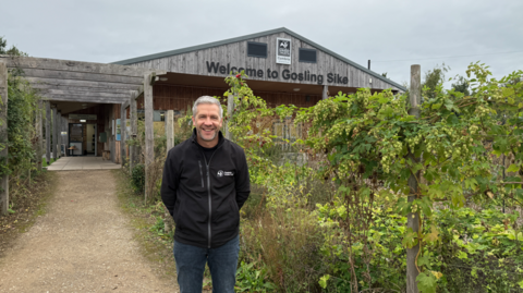 Adam McGinley is smiling in front of a path leading to Gosling Sike's entrance.  there are grape vines next to him. Adam has short white hair and is wearing jeans and a grey zip-up sweatshirt. 