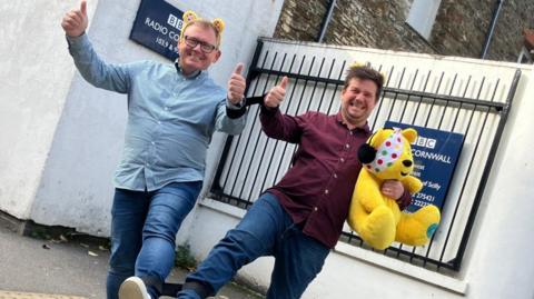 BBC Radio Cornwall presenters James Churchfield and James Dundon give thumbs up outside the radio station while their legs are tied together. Both have  Pudsey ears on their heads. James Dundon is holding a Pudsey teddy bear.