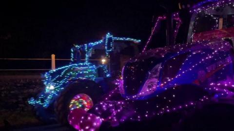 A night-time photo of two tractors covered in multi-coloured Christmas lights, with an agricultural fence in the background. 