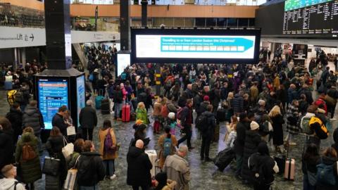 Passengers wait on a concourse beneath departure screens in London Euston station.