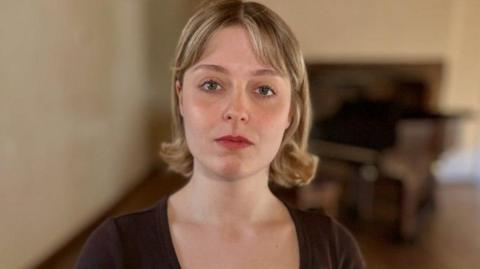 Lizzie Blissett looks directly at the camera. She is wearing a black scoop-neck dress and has her blonde hair in a bob. Behind her, blurred, is a piano.