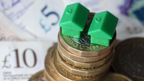 A stack of £1 coins with two small, green plastic houses sitting on top of them. The coins are on £5 and £10 notes.