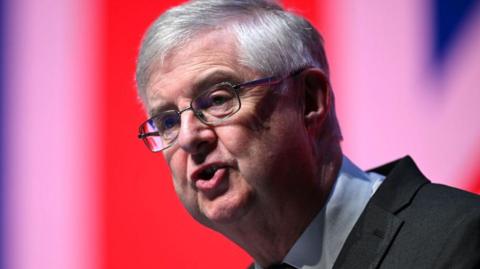 Mark Drakeford speaking while looking off camera to the left of the frame. He is swearing a suit and shirt, and glasses.