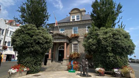 A picture of the outside of the proposed Victor Hugo Centre in Guernsey. It is a large brown building with a statue and flag outside.