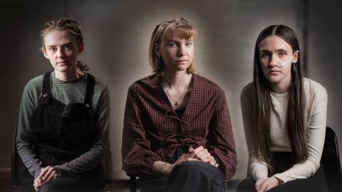 Three teenage girls in front of a grey background, one dressed in dark clothing, one in red, checked shirt and one in a red top with long dark hair.