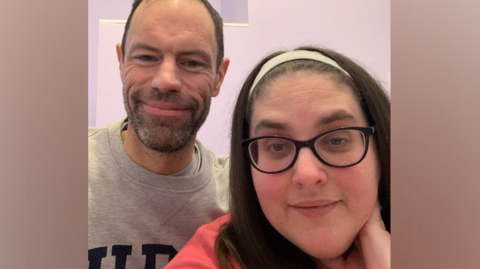 The image shows Alison Wright smiling directly at the camera. She is wearing glasses and has long, dark hair. Behind her, and also smiling at the camera, is her husband James. He has a dark beard and short hair.