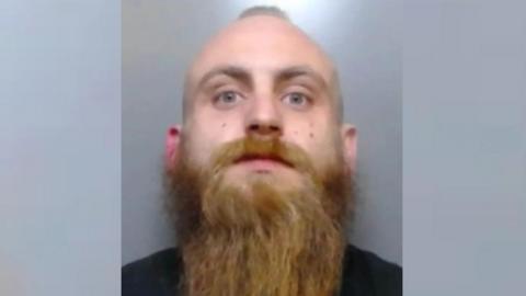 Mugshot of a man with a little hair in the middle of his head and a long brown beard staring at the camera. He is wearing a black shirt.