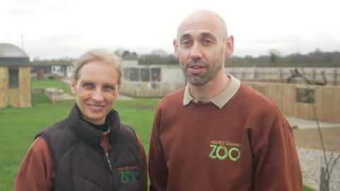 A blonde-haired woman in a black gilet stands next to a man in a brown "Holmes Chapel Zoo" branded jumper. Behind them are wooden enclosures.