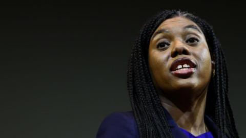 Kemi Badenoch wearing blue outfit against plain dark backdrop