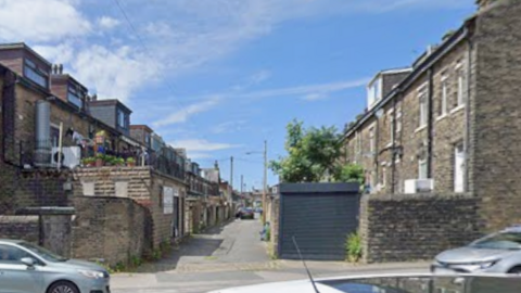 View of a narrow alleyway between rows of stone terraced houses in Bradford, with parked cars on either side of the entrance.