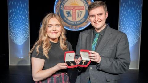 Alice MacMillan and Ryan Johnston showing their medals, which are in red presentation boxes. The Royal National Mòd logo is in the background.