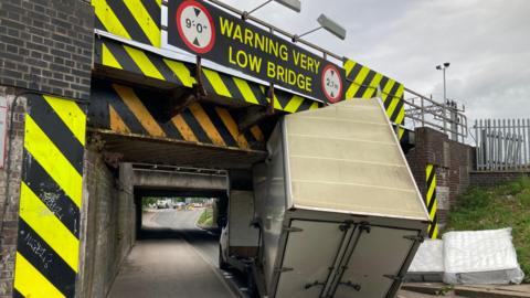 The rear of a white van has struck a railway bridge which a single track road runs below. There is a large yellow and black sign that says WARNING VERY LOW BRIDGE above the road.