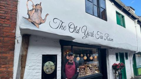 Owner Tony Vickers standing outside The Old York Tea Room, where a tea pot and the tea room's name is painted on the building 