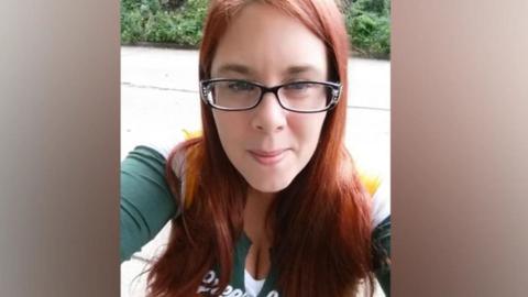 A woman with red hair, dark-rimmed glasses and a green and white top is looking towards a camera. There is greenery in the background.
