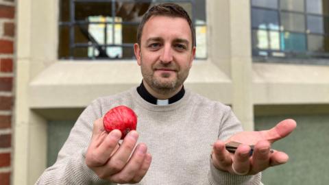 Mr Pearce is wearing a beige jumper, but his clerical collar can be seen around his neck. Using both hands, he is holding up a red apple and shards of glass to the camera. Behind him are the church's stained glass windows. Mr Pearce has short brown hair and stubble.