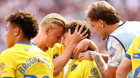 Footballers Adrian Segecic and Rocco Robert Shein grinning at each other while locking heads to celebrate a Portsmouth Football Club goal during their Carabao Cup first round match against West Ham United at London Stadium while wearing a predominantly yellow kit with blue details and surrounded by teammates