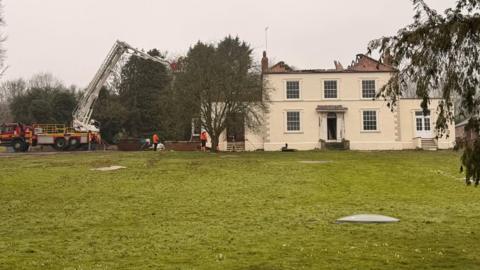 A large detached house with cream-coloured rendered walls. The roof appears to be badly damaged with just a small section to the right of the image being intact. There is a fire engine with an aerial platform to the side.