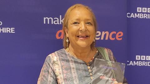 Linda, a woman with short golden hair, wearing a grey top, standing in front of a purple BBC Cambridgeshire Make a Difference board, holding a glass award.