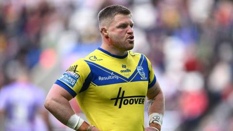 Luke Yates in action for Warrington. Seen only from the waist up Yates is standing with his hands on his hips, wearing a Warrington home jersey in the traditional yellow and blue. He has blood trickling down his jaw from his left ear.