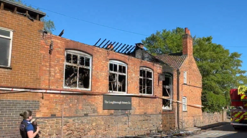 A brick building with extensive damage to its roof and windows. A police officer stands outside and a fire engine can be seen
