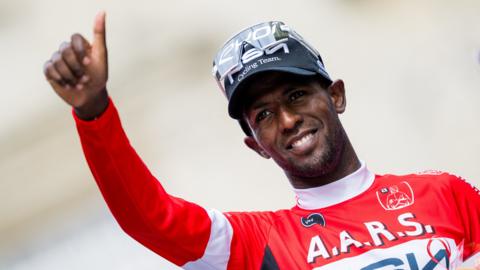 Biniam Girmay, wearing a red long-sleeved cycling jersey and clear sunglasses on top of a blue baseball cap, smiles as gives a thumbs up gesture with his right arm raised in front of him