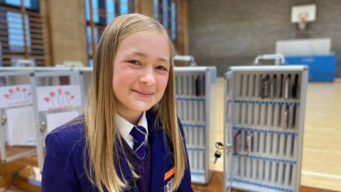 Mila is in her school uniform, smiling to camera, behind her are mobile phone storage lockers where students can store their smartphones safely during the school day