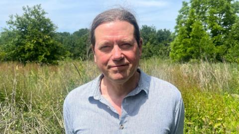 A man standing in a rewilded garden. He is smiling at the camera while wearing a baby blue shirt 