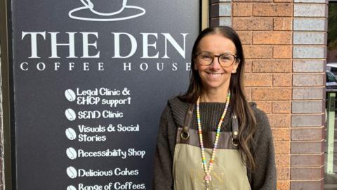 Claire Naylor is wearing a black jumper with an apron, and glasses, standing in front of a large sign for The Den Coffee House which lists its services. Services include a Send clinic, an accessibility shop and support with education, health and care plans.