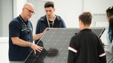 A Hull College staff member gestures towards a solar panel as a group of older teenagers watch