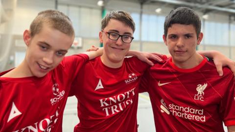 Alife, MacKenzie, and Callum are wearing matching red football shirts stand together inside an indoor sports hall, each with an arm around the shoulders of the person next to them. The shirts feature logos including “Knott Trust” and “Standard Chartered.” The sports hall has court markings and a high industrial-style ceiling.