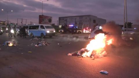 A pile of rubbish burns on the highway at dusk as traffic tries to get past.