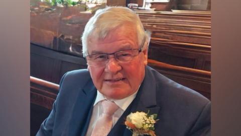 A man wearing a navy blue suit along with a white shirt and a pink tie is sitting on a church pew. He has a flower in the lapel of his suit jacket. He has grey hair and is wearing glasses.